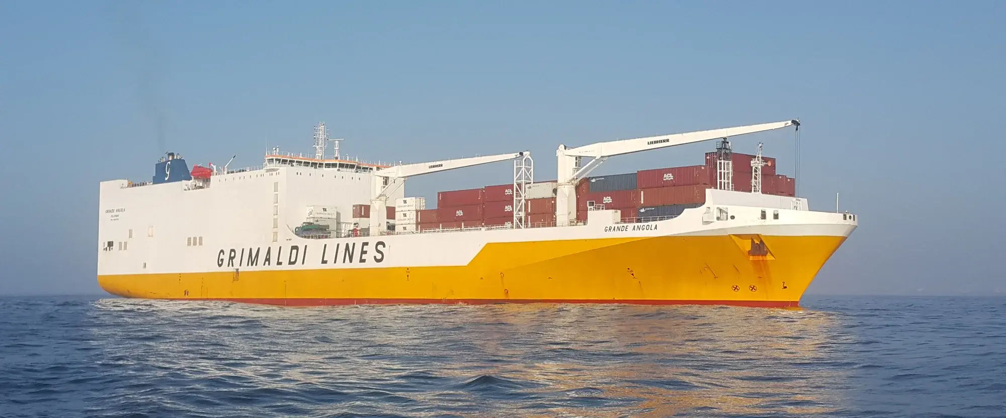 The Grimaldi Lines car carrier Grande Angola at sea under a clear sky, containers on her after deck and GRIMALDI LINES along the white hull